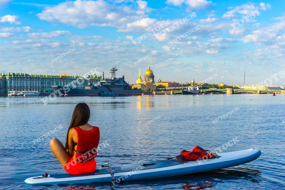 На фотографии изображена девушка на сапборде, отдыхающая на Неве с видом на знаменитые достопримечательности Санкт-Петербурга — Исаакиевский собор и Зимний дворец. Летний день с ясным небом и спокойной водой подчёркивает красоту исторического центра города. На заднем плане видны также корабли, создавая впечатляющий контраст между современной активностью и классической архитектурой города.